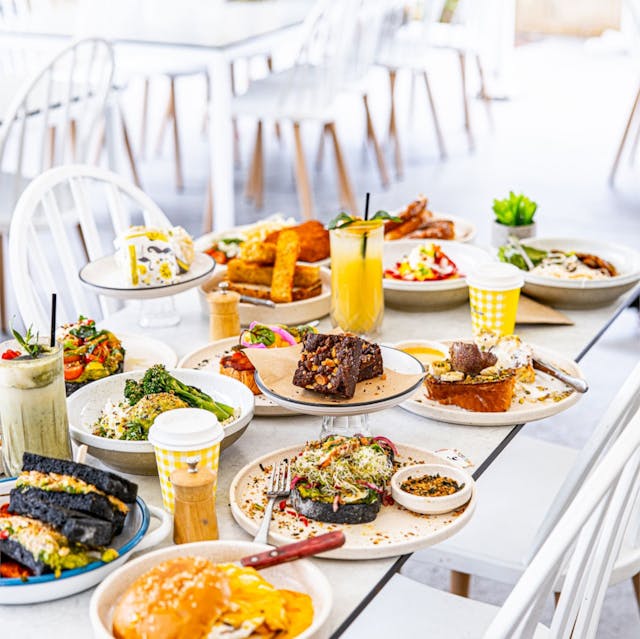 An array of dishes displayed on a white table in a bright dining setting. The spread includes various beverages, salads, grilled items, desserts, and sandwiches, all presented on assorted plates and glasses